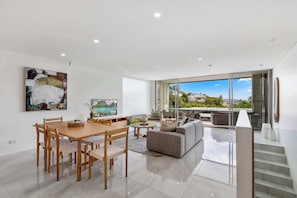A modern living room with a dining area, a sofa, and a large window offering a view of the outdoors. - Contemporary Townhouse, Heated Pool, Walk to Beach (Noosa Region)