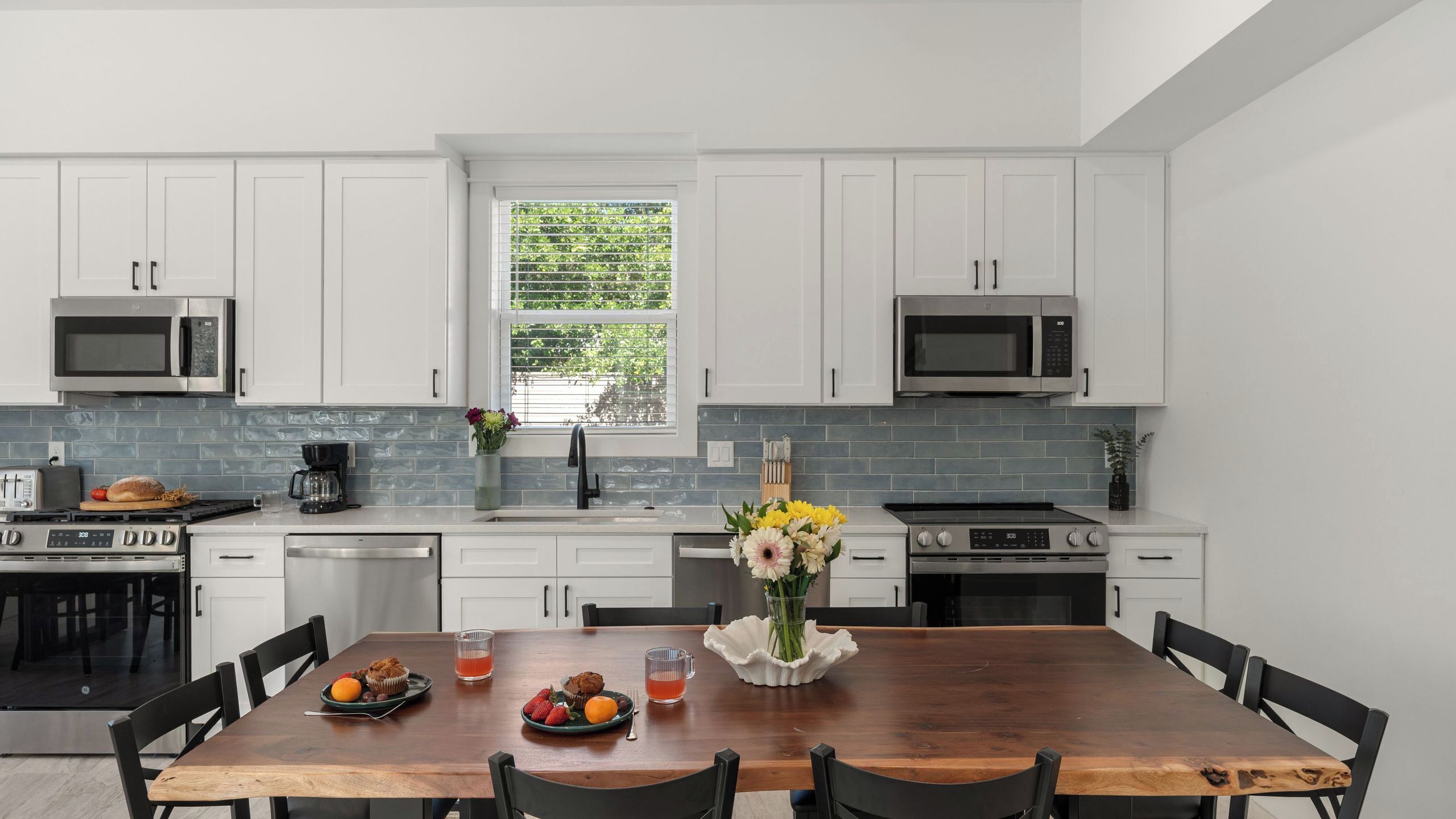 A modern kitchen with white cabinets, two stainless steel microwaves, a built-in oven, and a dining table with chairs.