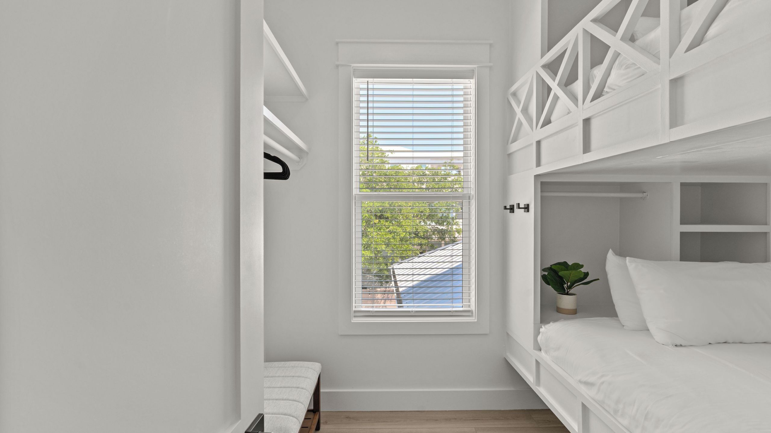 Ein Zimmer mit heller Einrichtung, einem Fenster mit Jalousien, einem Bett und einer kleinen Pflanze.