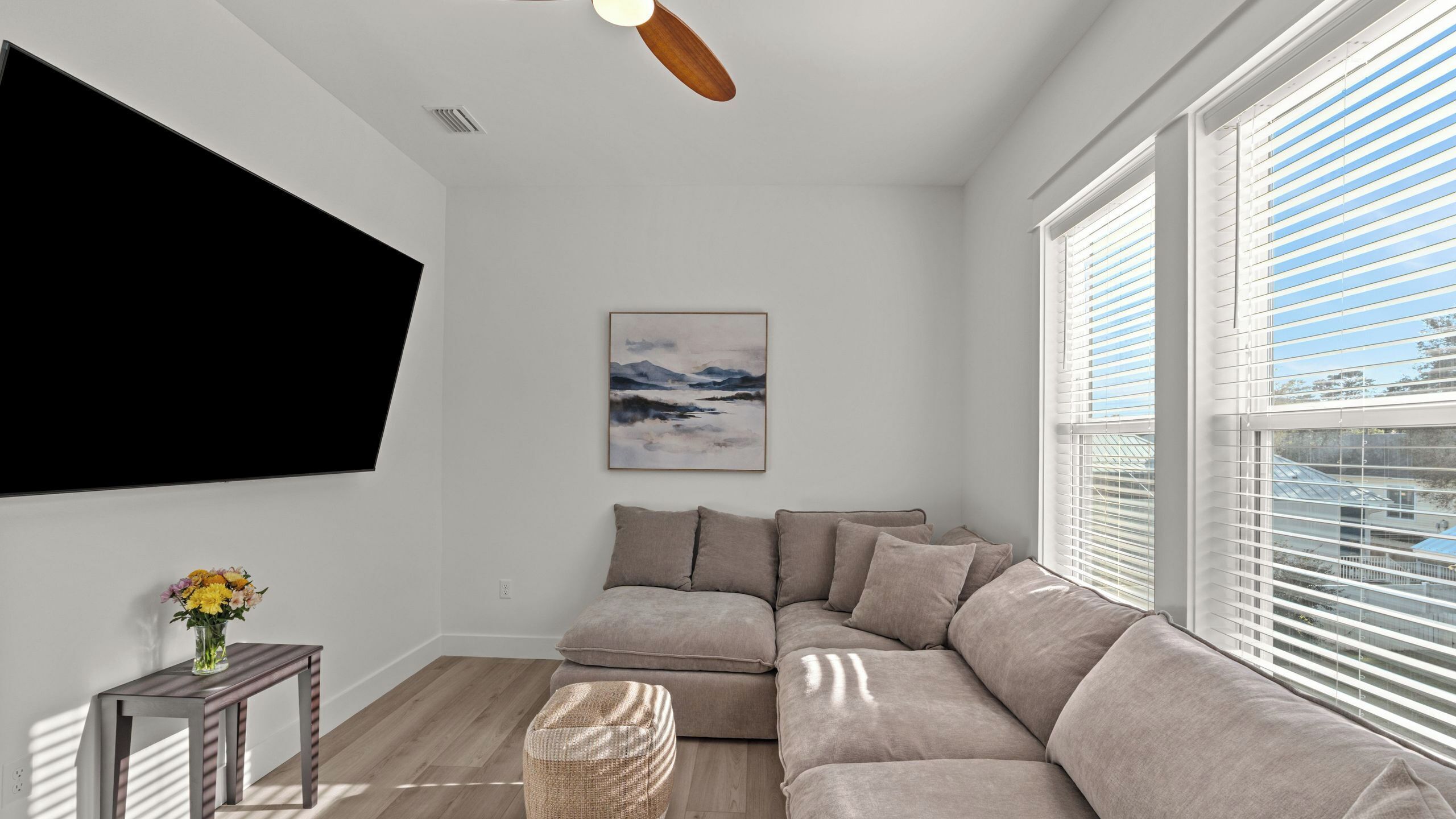 Ein Wohnzimmer mit einem Ecksofa, einem flachbildfernseher an der Wand, einem Deckenventilator und einem Fenster mit Jalousien.