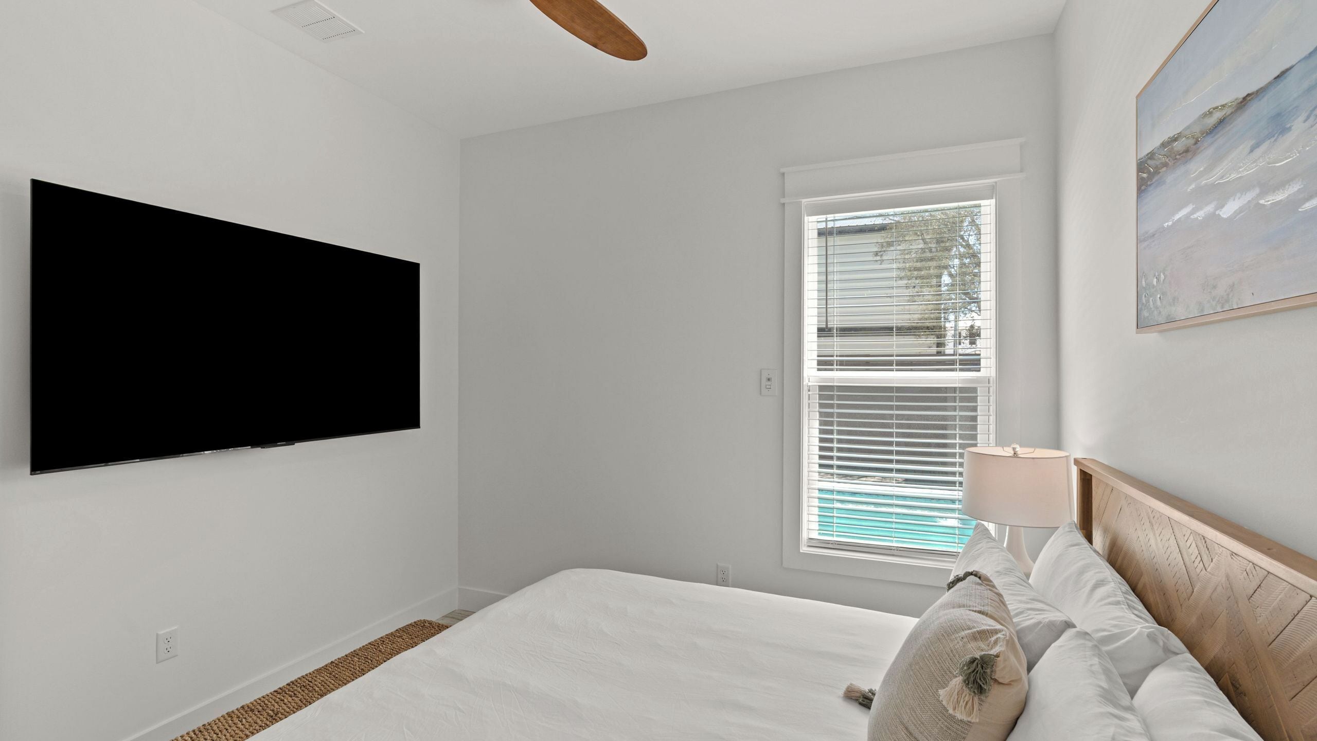 A bedroom with a bed, a TV mounted on the wall, a window with blinds, and a painting on the wall.