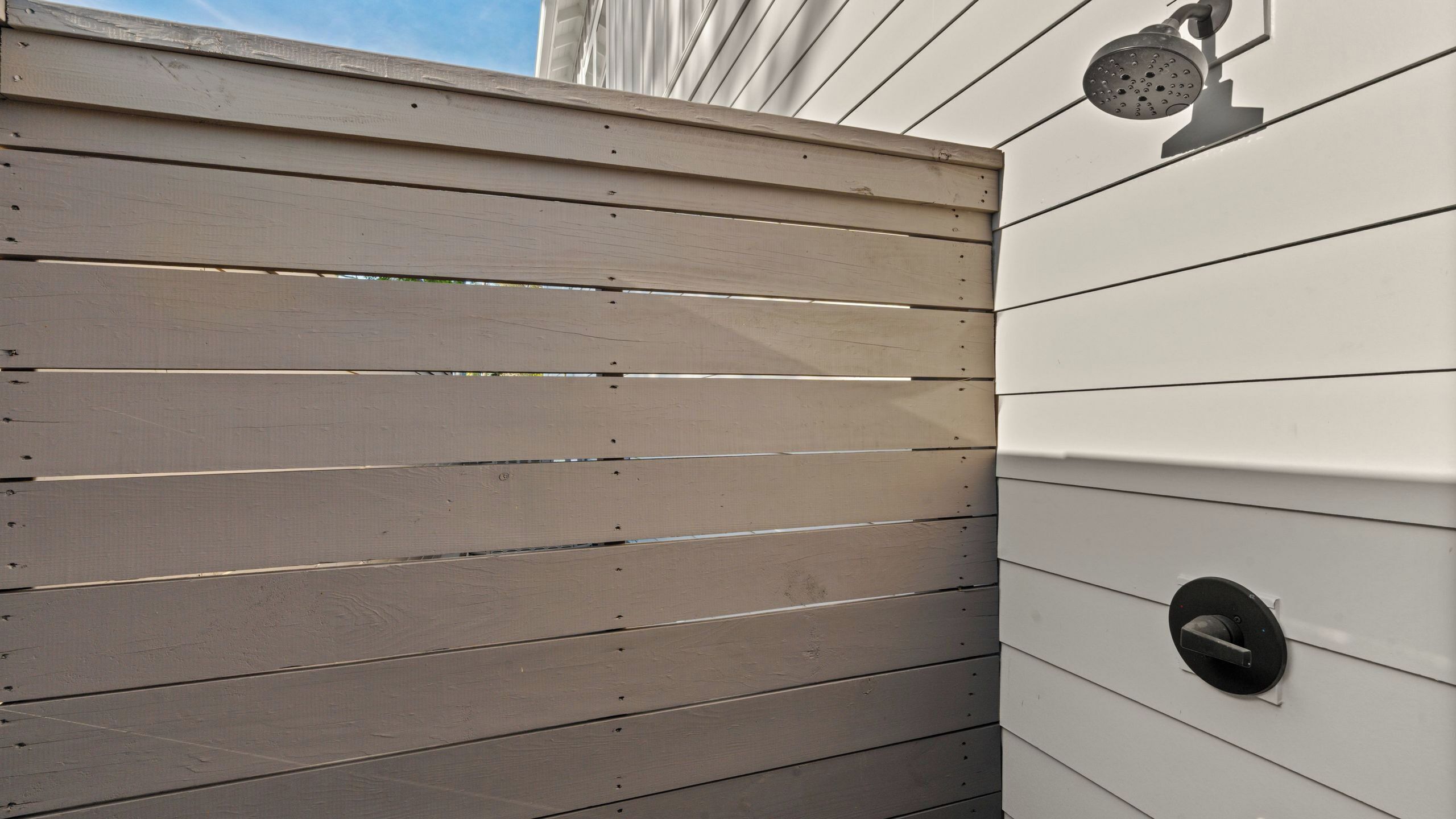 Une clôture en bois avec des lattes horizontales, une porte en métal dotée d’une poignée ronde et une pomme de douche en métal fixée à un mur blanc.