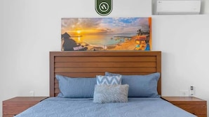 Un dormitorio con cabecera de madera, cama con colcha azul estampada, dos almohadas y un cuadro enmarcados con una escena de playa sobre la cama.