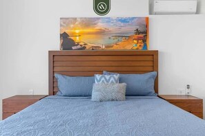 Ein Schlafzimmer mit einem Holzkopfteil, einem Bett mit einer blauen, gemusterten Tagesdecke, zwei Kissen und einem über dem Bett hängenden Bild von einer Strandansicht.