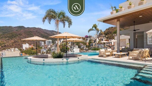 Zona de piscina con sillones de descanso y sombrillas, rodeada de edificios y palmeras.