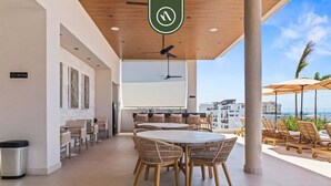 Amplio espacio interior con techo alto, ventilador de techo y una pantalla grande. Varias mesas y sillas están dispuestas para comer o socializar. El ambiente está bien iluminado gracias a la luz natural que entra por grandes ventanales.