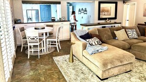 Un salon moderne avec un canapé d’angle, un coin repas avec une table et des chaises blanches, et une cuisine avec des meubles bleus et un réfrigérateur.