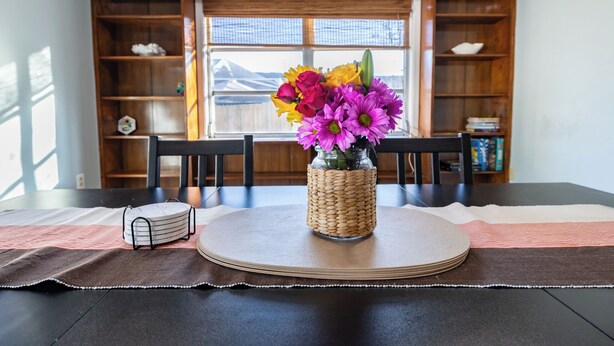 Une table à manger ornée d’un vase tressé rempli de fleurs, d’un set de table et d’une petite pile de livres.