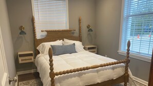 A bedroom with a wooden four-poster bed, a nightstand, a wall-mounted lamp, and a window with blinds. - The Nook on Main St. (Hudson)