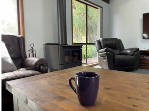 A cozy living room with a fireplace, leather chairs, a wooden coffee table, and a large window with a view of trees.