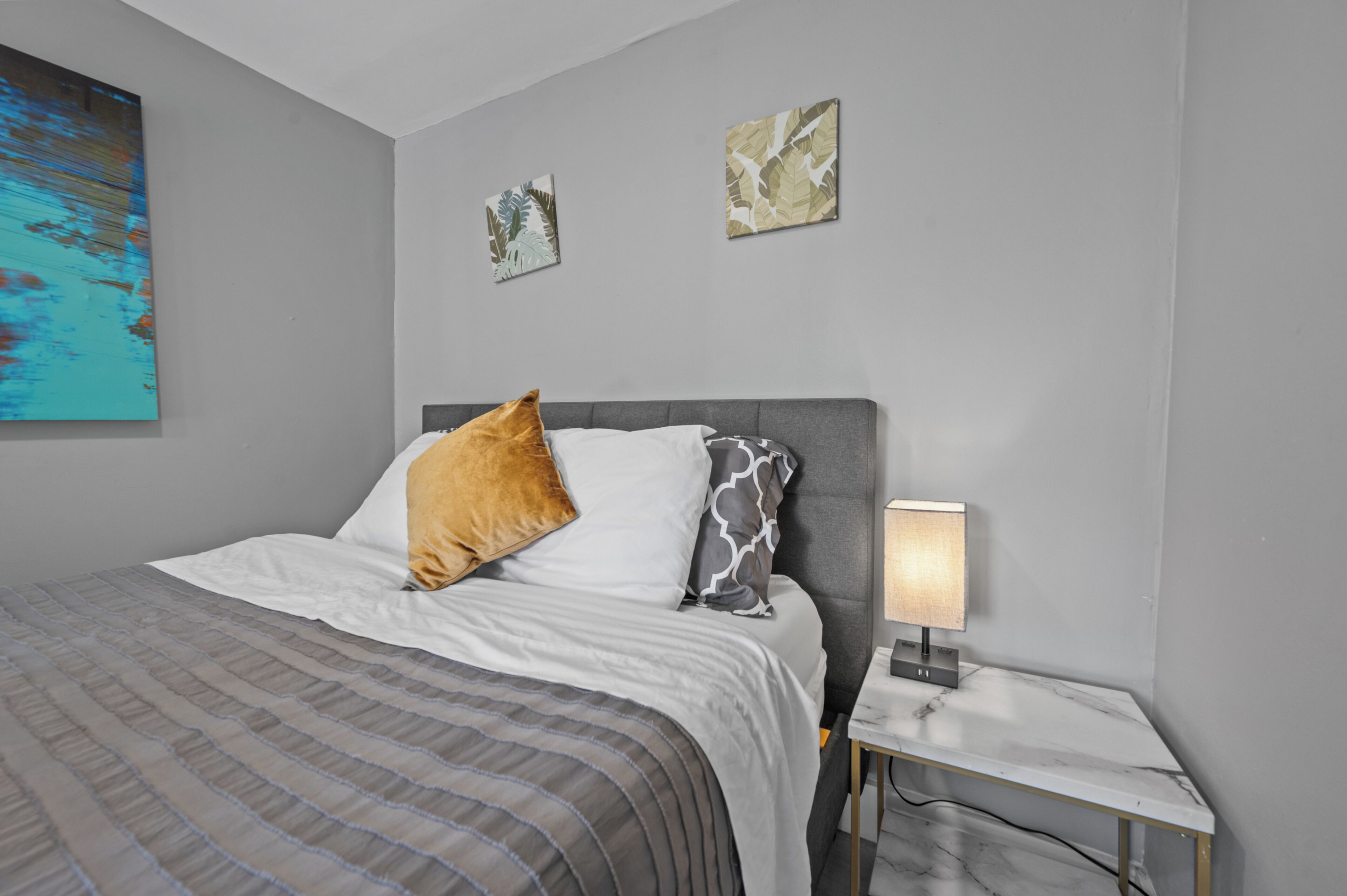 Un lit bien fait, recouvert d’une couvre-lit à rayures, une table de chevet avec une lampe, et deux œuvres d’art accrochées au mur.