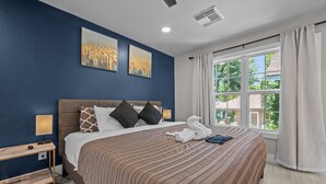 A bedroom with a large bed, a nightstand, two wall-mounted paintings, a window with curtains, and a view of greenery outside.