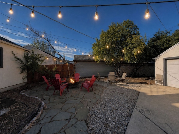 A backyard with stone flooring, a fire pit, red chairs, and string lights.