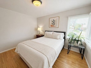 A bedroom with a bed, bedside table, lamp, framed artwork, and a window with blinds.