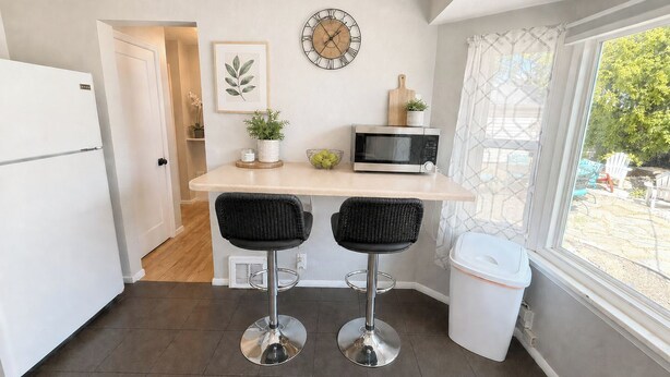 A compact kitchen with a microwave, two bar stools, a clock, and a window with a view of greenery. - Sugarhouse Retreat - Walk to Shops + Parks - Cozy Home w/ Private Yard (Salt Lake City)
