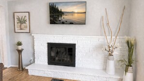 A living room with a white brick fireplace, a framed landscape painting, and two white vases with plants. - Sugarhouse Retreat - Walk to Shops + Parks - Cozy Home w/ Private Yard (Salt Lake City)
