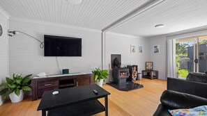 A modern living room with a flat-screen TV, a fireplace, wooden furniture, and a view of the outdoors. - Seabreeze Cottage Pet Friendly Family Holiday Home close to the Beach (Manyana)
