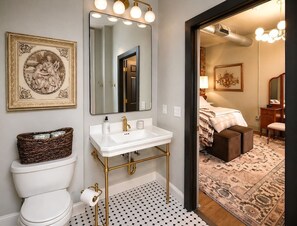 Een badkamer met een wit toilet, een witte wastafel met gouden details, een grote spiegel, en een kamerbreed vloerkleed met een patroon in de aangrenzende slaapkamer.