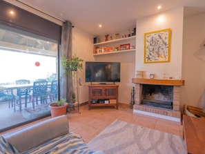 A living room with a TV, fireplace, and outdoor seating visible through a glass door. - Can Jamy by Costabravaway (calonge)