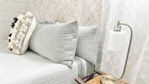 A bed with a quilted cover, two pillows, a woven side table with a lamp, and a headboard with a herringbone pattern. - 253 Upper Chic Boho Retreat NEW RENO (Erie)