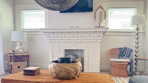 A living room with a white brick fireplace, a wooden table with a woven basket, a wicker chair, and a lamp. - 253 Upper Chic Boho Retreat NEW RENO (Erie)