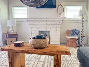A living room with a white brick fireplace, a wooden table with a woven basket, a wicker chair, and a lamp. - 253 Upper Chic Boho Retreat NEW RENO (Erie)