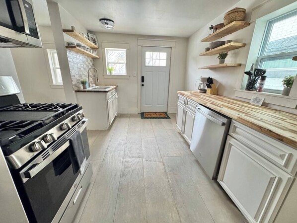 A kitchen with a stove, oven, and white cabinets. There are wooden shelves above the counter and a window with a view. - Renting Erie Property. 4br, living room, dining room, laundry, dishwasher, Only downside- just 1 bathroom. (Erie)