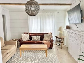 A living room with a brown leather sofa, a wooden coffee table, a patterned rug, a white brick fireplace, and a woven light fixture.