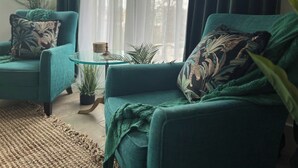 En soffa i en nyans av turkos med ett mönstrat kuddöverdrag, ett soffbord i glas och en vävd matta i ett vardagsrum med utsikt över träd genom tunna, genomskinliga gardiner.