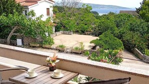 Un balcón con mobiliario de mimbre, una mesa con un florero y tazas, y vistas a una casa y follaje.