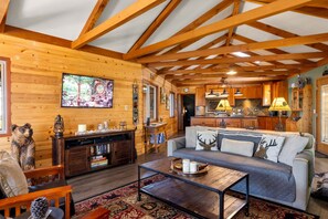 A log cabin interior with a living room, kitchen, and dining area, featuring wooden beams, a sofa, and a television.