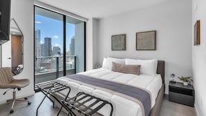 A modern bedroom with a large bed, a desk with a chair, a view of the city through a balcony, and two abstract wall art pieces. - Modern High Rise Stay- 23 Floor Views (Miami)