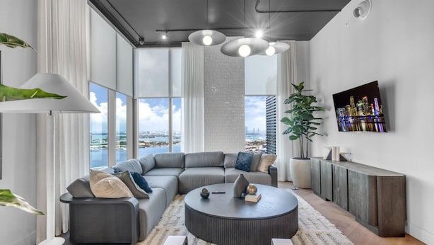 A modern living room with a sectional sofa, a round coffee table, a TV mounted on the wall, and large windows offering a city view. - Modern High Rise Stay- 23 Floor Views (Miami)