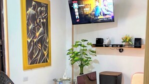A room with a ceiling fan, a flat-screen TV mounted on the wall, a laptop on a mobile desk, a potted plant, a black chair, and a wooden shelf with speakers.