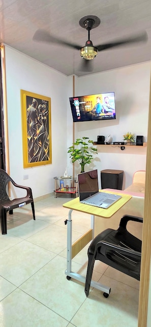 A room with a ceiling fan, a flat-screen TV mounted on the wall, a laptop on a mobile desk, a potted plant, a black chair, and a wooden shelf with speakers. - Suitable for 7 very close to Cafam (Melgar)