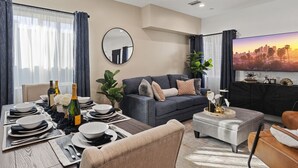 Un salon moderne avec un coin repas aménagé pour un repas, un canapé gris, un téléviseur et une vue sur le paysage urbain.