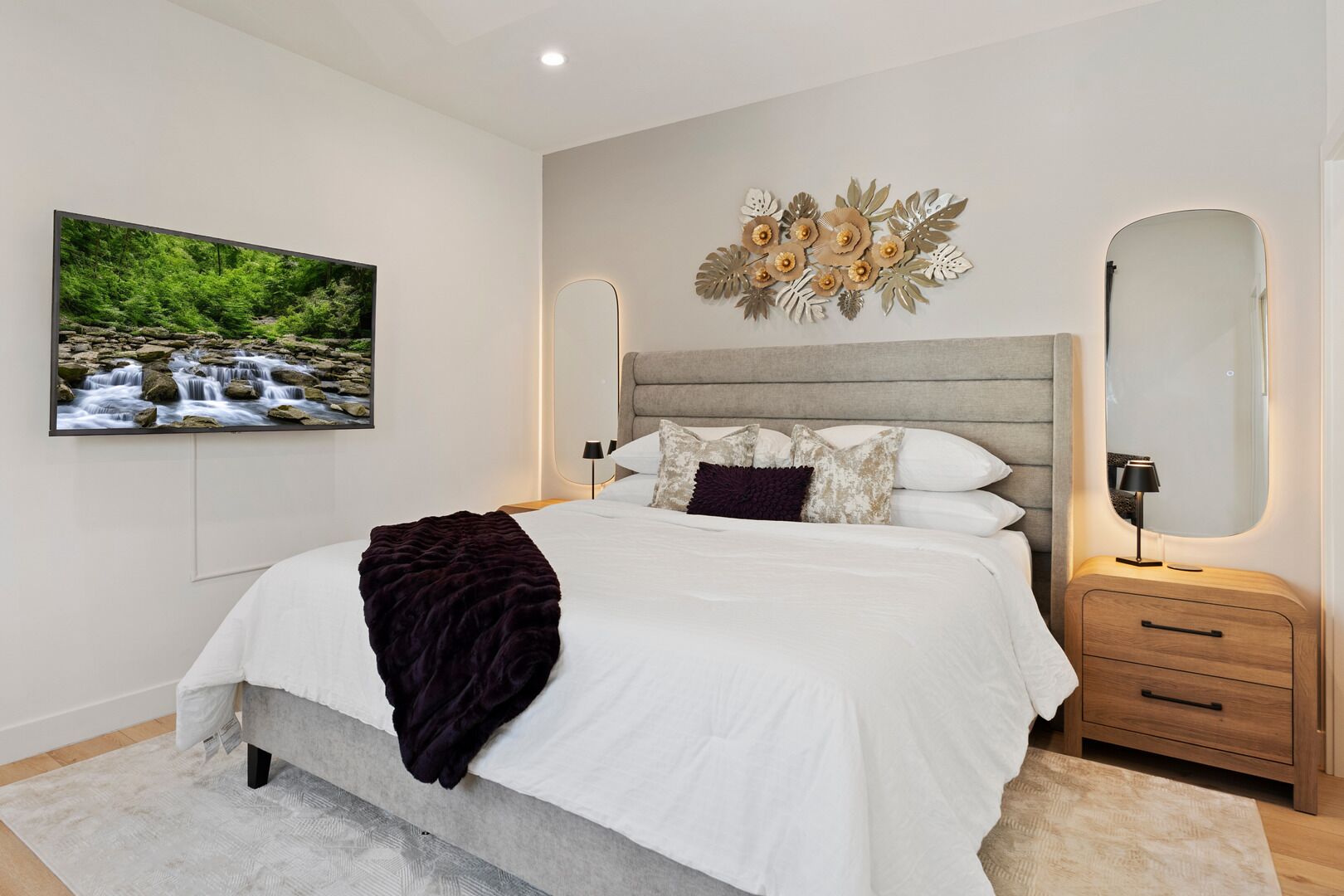 A modern bedroom with a large bed, a headboard with a decorative floral design, a bedside table with a lamp, and a framed picture of a waterfall on the wall.
