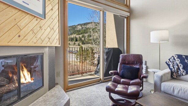 A room with a fireplace, a leather chair, a sofa, a lamp, and a view of a forest through a sliding glass door. - She305 Summit House East 1 Bedroom Condo (Copper Mountain)