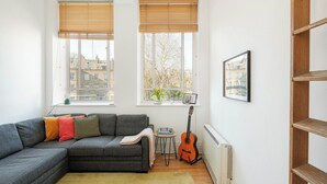 A modern living room with a grey sectional sofa, a small table, a guitar, and a radiator. - Bright 1BR in Central Clerkenwell (London)