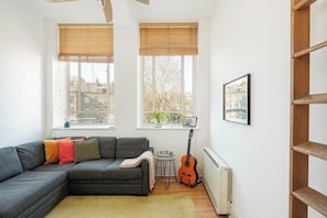 A modern living room with a grey sectional sofa, a small table, a guitar, and a radiator. - Bright 1BR in Central Clerkenwell (London)