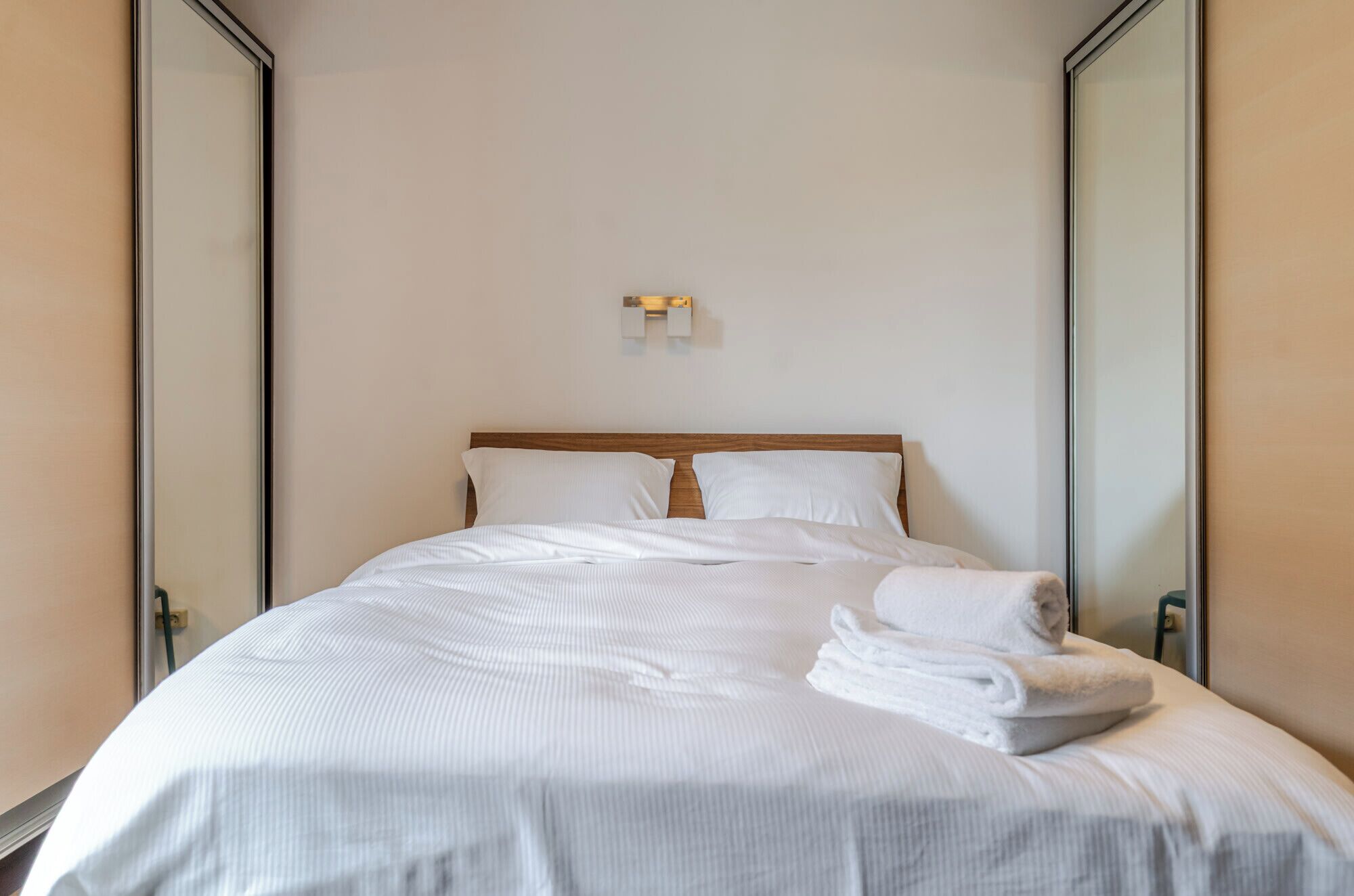 Un lit bien fait, avec du linge de lit blanc, une tête de lit en bois et un petit luminaire fixé au mur.
