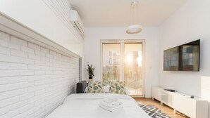 A modern bedroom with white brick walls, a bed with white bedding, a patterned rug, a side table, and a mounted TV. - Rakowicka Charming Apartment with Garden (Kraków)