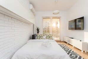 Ein modernes Schlafzimmer mit weißen Ziegelwänden, einem Bett mit weißer Bettwäsche, einem gemusterten Teppich, einem Nachttisch und einem angebrachten Fernseher.