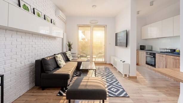 A modern living room with a sofa, ottoman, and a dining table. There is a kitchen area with cabinets and a TV mounted on the wall. - Rakowicka Charming Apartment with Garden (Kraków)
