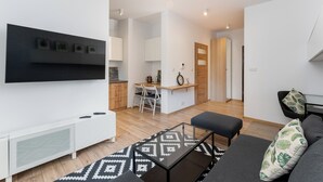 A modern living room with a sofa, ottoman, and a coffee table. There is a kitchen area with a dining table and chairs, and a small island with a clock. - Rakowicka Charming Apartment with Garden (Kraków)