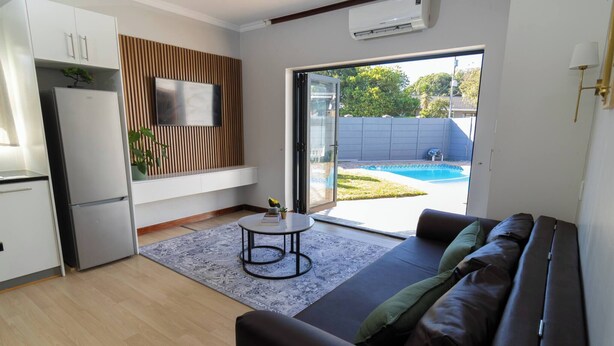 Un soggiorno moderno con divano, un tavolino, una TV montata su un pannello di legno e una porta scorrevole in vetro che dà su una zona esterna con piscina.