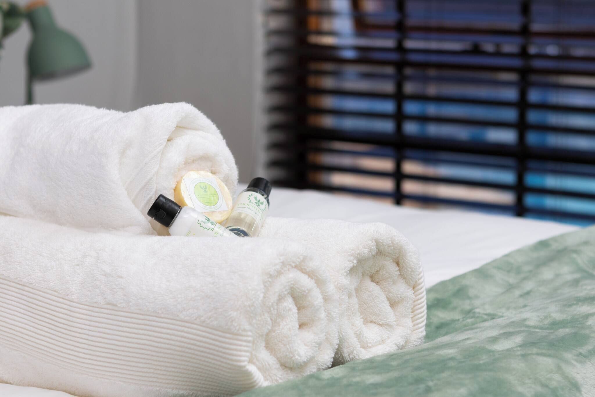 Een keurig opgevouwen witte handdoek op een bed met een groene geborduurde sprei, een flesje lotion en een klein flesje shampoo.