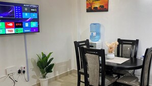 A modern living room with a flat-screen TV, a dining table with chairs, a potted plant, and a water dispenser.