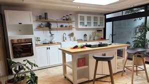 Une cuisine moderne avec des meubles blancs, un îlot central doté de tabourets, un four encastré et une grande fenêtre qui laisse entrer la lumière naturelle.