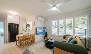 A modern living room with a dining area, a black refrigerator, a flat-screen TV, a ceiling fan, and large windows with blinds. - KozyGuru | Mosman | Serene Boutique Apt Retreat (Mosman)